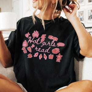 Może przedstawiać: Czarny t-shirt z różowym napisem "Hot girls read", otoczony różowymi ilustracjami: kubkiem, okularami, błyskawicą i książką. Koszulka ma swobodny styl.