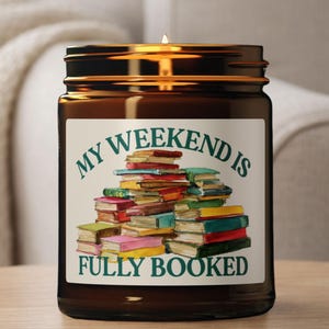 May include: A brown glass jar candle with a lit wick. The label reads "MY WEEKEND IS FULLY BOOKED" above a colorful stack of books. The candle is a decorative item for book lovers.