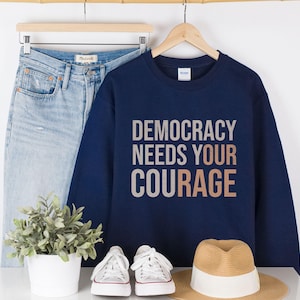 Puede incluir: Una sudadera azul marino con el texto "DEMOCRACY NEEDS YOUR COURAGE" en letras blancas y cobrizas. La sudadera se muestra con unos vaqueros azul claro, zapatillas blancas y un sombrero de paja.