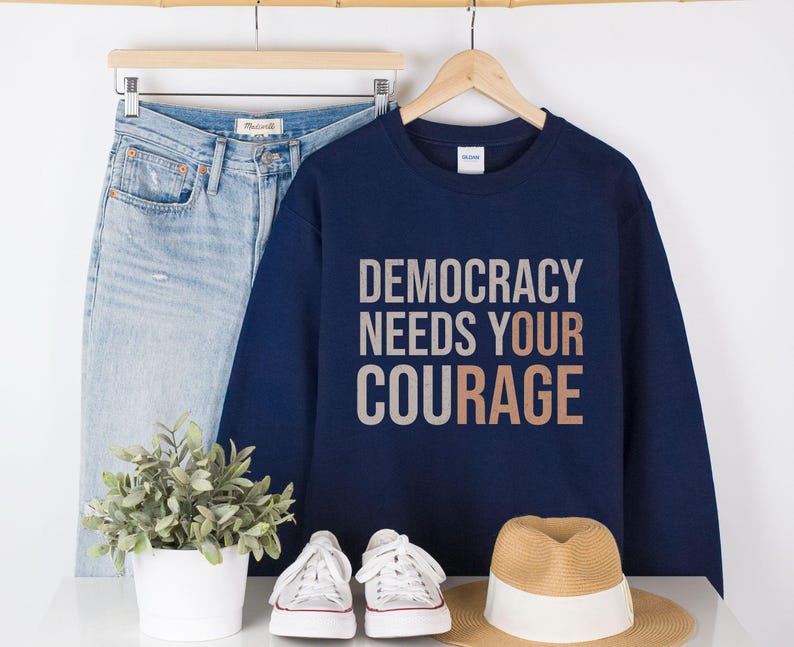 Puede incluir: Una sudadera azul marino con el texto "DEMOCRACY NEEDS YOUR COURAGE" en letras blancas y cobrizas. La sudadera se muestra con unos vaqueros azul claro, zapatillas blancas y un sombrero de paja.