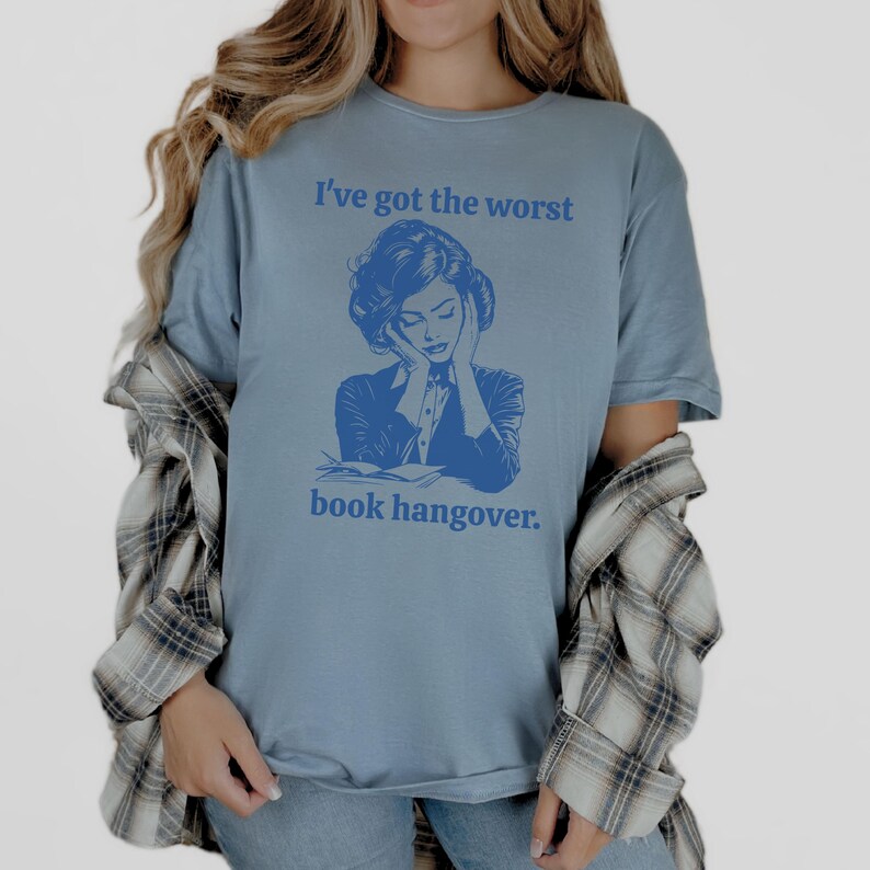 Op de afbeelding: Een blauw t-shirt met een afbeelding van een vrouw die haar hoofd in haar handen houdt en de tekst "Ik heb de ergste boeken kater."