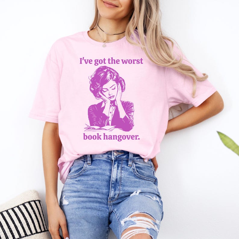 Op de afbeelding: Een roze T-shirt met een afbeelding van een vrouw die haar hoofd in haar handen houdt en de tekst "Ik heb de ergste kater van een boek."