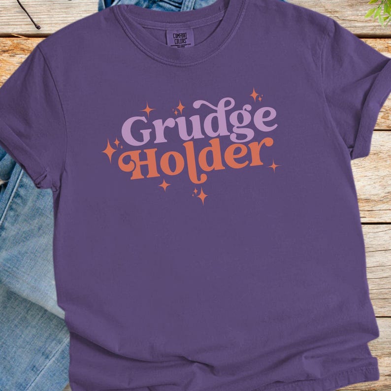 May include: Purple t-shirt with the text "Grudge Holder" in pink and orange letters with sparkling stars around the text.