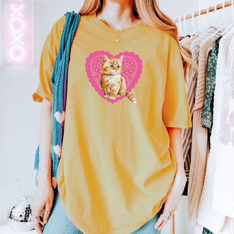 May include: A yellow oversized t-shirt with a pink heart-shaped design featuring a cartoon orange tabby cat.