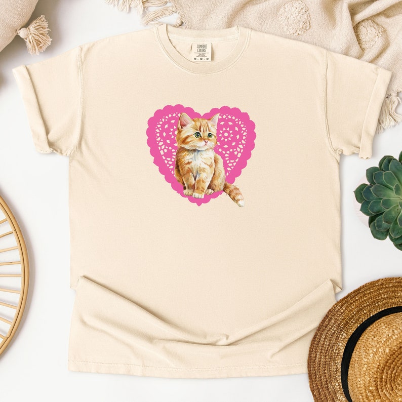 May include: A cream colored t-shirt with a pink heart-shaped doily featuring a cartoon orange tabby kitten.