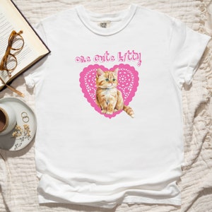 Op de afbeelding: Wit T-shirt met een roze hartvormig kantontwerp en een schattig oranje kitten erin. De tekst "one cute kitty" is boven het hart gedrukt.