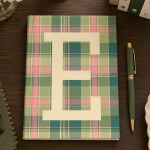 May include: A notebook with a green, pink, and white plaid design and a large cream-coloured letter "E". A green pen with gold detailing lies next to the notebook. The notebook is on a dark wood surface.