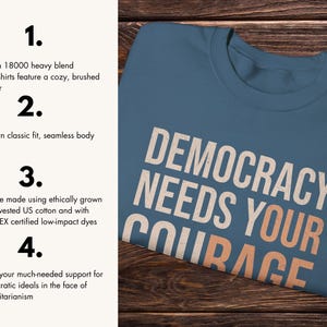 Puede incluir: Una camiseta azul con la frase "DEMOCRACY NEEDS YOUR COURAGE" en letras blancas y naranjas. La camiseta est&aacute; hecha de algod&oacute;n estadounidense cultivado y cosechado &eacute;ticamente.