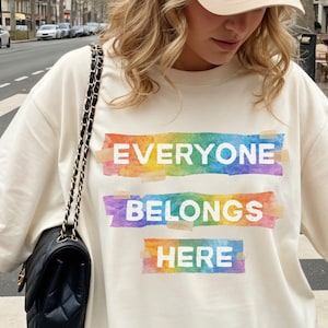 Może przedstawiać: Kremowa koszulka z napisem "EVERYONE BELONGS HERE" w białych literach. Tekst jest nałożony na trzy poziome tęczowe paski w stylu akwareli. Koszulka ma luźny krój.