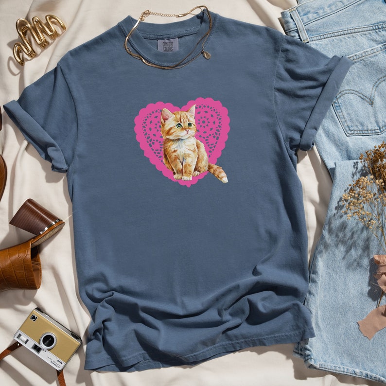 May include: A blue t-shirt with a pink heart-shaped design featuring a cartoon orange tabby kitten.
