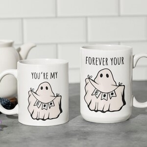 May include: Two white ceramic mugs with black text and ghost illustrations. One mug reads "YOU'RE MY BOO" and the other reads "FOREVER YOUR BOO". The ghost design includes a banner with the word "BOO".
