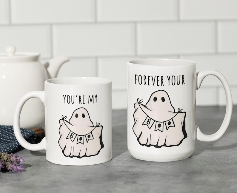 May include: Two white ceramic mugs with black text and ghost illustrations. One mug reads "YOU'RE MY BOO" and the other reads "FOREVER YOUR BOO". The ghost design includes a banner with the word "BOO".