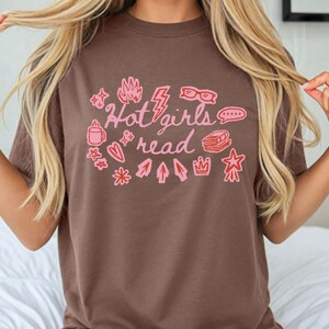 May include: Brown t-shirt with the text "Hot girls read" in pink, surrounded by pink illustrations of a coffee mug, stars, a flame, glasses, a lightning bolt, a speech bubble, a book, a crown, and arrows.