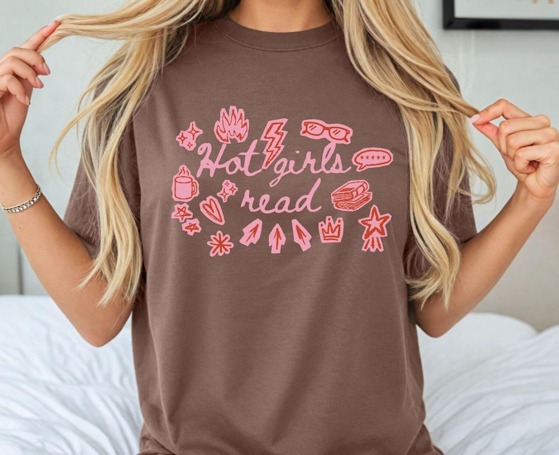 May include: Brown t-shirt with the text "Hot girls read" in pink, surrounded by pink illustrations of a coffee mug, stars, a flame, glasses, a lightning bolt, a speech bubble, a book, a crown, and arrows.