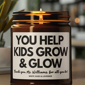May include: A lit brown glass jar candle with a white label. The label reads "YOU HELP KIDS GROW & GLOW" with the text "Thank you, Ms. Williams, for all you do!" and "WHITE SAGE & LAVENDER". The candle is in front of a blurred background.
