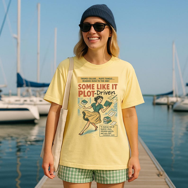 May include: A pale yellow t-shirt featuring the text "SOME LIKE IT PLOT-DRIVEN" and a retro illustration of a woman with books. The shirt also has text about plots and readers. The person is wearing a blue beanie and sunglasses.