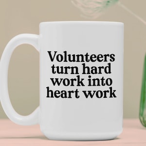 Pode incluir: Caneca de cerâmica branca com uma alça grande. A caneca apresenta o texto "Volunteers turn hard work into heart work" em preto, em negrito. A caneca está sobre uma superfície clara, com um vaso verde no fundo.