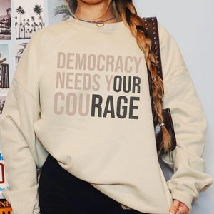 Puede incluir: Sudadera color crema con el texto "DEMOCRACY NEEDS YOUR COURAGE" en tonos grises. La sudadera tiene mangas largas y cuello redondo. El fondo incluye im&aacute;genes de palmeras y un letrero de "NO".