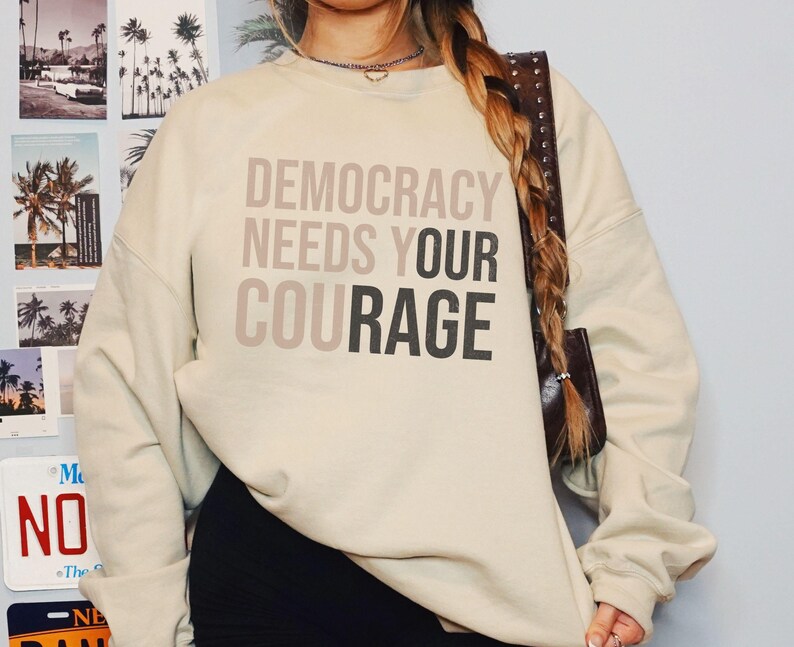 Puede incluir: Sudadera color crema con el texto "DEMOCRACY NEEDS YOUR COURAGE" en tonos grises. La sudadera tiene mangas largas y cuello redondo. El fondo incluye im&aacute;genes de palmeras y un letrero de "NO".