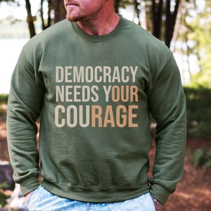 Puede incluir: Sudadera verde oliva con el texto "DEMOCRACY NEEDS YOUR COURAGE" en una fuente de color neutro en capas. La sudadera de manga larga parece estar hecha de un material suave. El fondo es una escena borrosa al aire libre.
