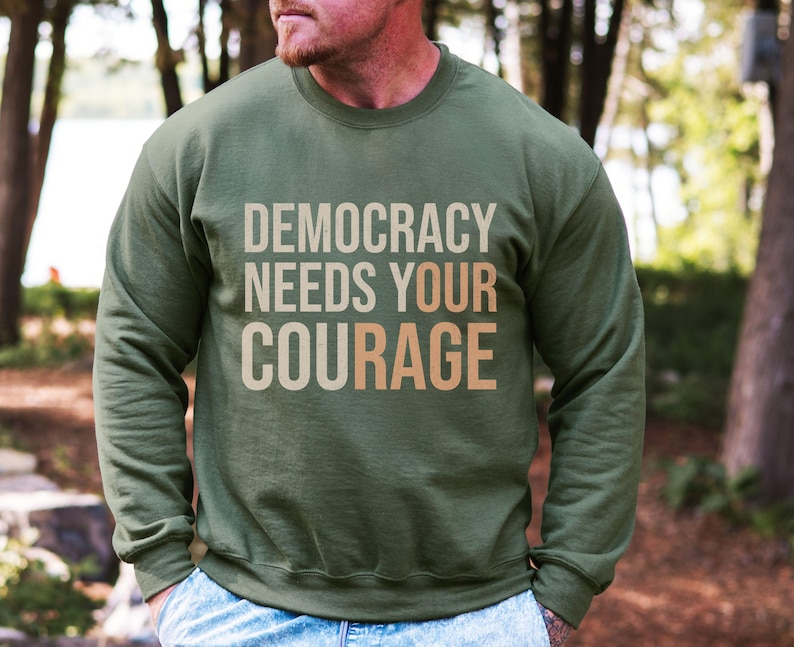 Puede incluir: Sudadera verde oliva con el texto "DEMOCRACY NEEDS YOUR COURAGE" en una fuente de color neutro en capas. La sudadera de manga larga parece estar hecha de un material suave. El fondo es una escena borrosa al aire libre.