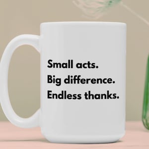 Pode incluir: Caneca de cerâmica branca com o texto "Small acts. Big difference. Endless thanks." em preto. A caneca tem uma alça grande e está sobre um fundo neutro. Um vaso verde é visível no fundo.