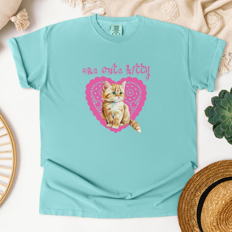 Op de afbeelding: Een lichtblauw T-shirt met een roze hartvormig ontwerp met een schattig oranje kitten. De tekst "one cute kitty!" is boven het hart gedrukt.