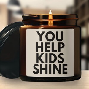 May include: A lit candle in a brown glass jar with a black lid. The jar has a white label with the words "YOU HELP KIDS SHINE" in bold black letters. The lid is off to the side.
