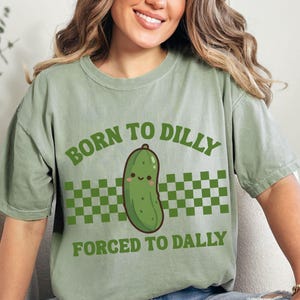 May include: Sage green t-shirt featuring the text "BORN TO DILLY FORCED TO DALLY" in green. A cartoon pickle with a smiling face is centered on the shirt, above a green and white checkered pattern.