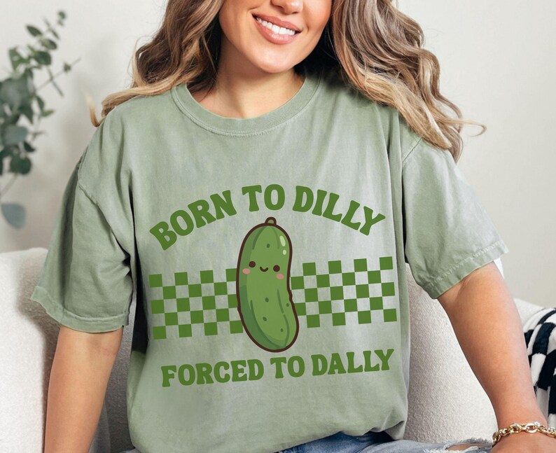 May include: Sage green t-shirt featuring the text "BORN TO DILLY FORCED TO DALLY" in green. A cartoon pickle with a smiling face is centered on the shirt, above a green and white checkered pattern.