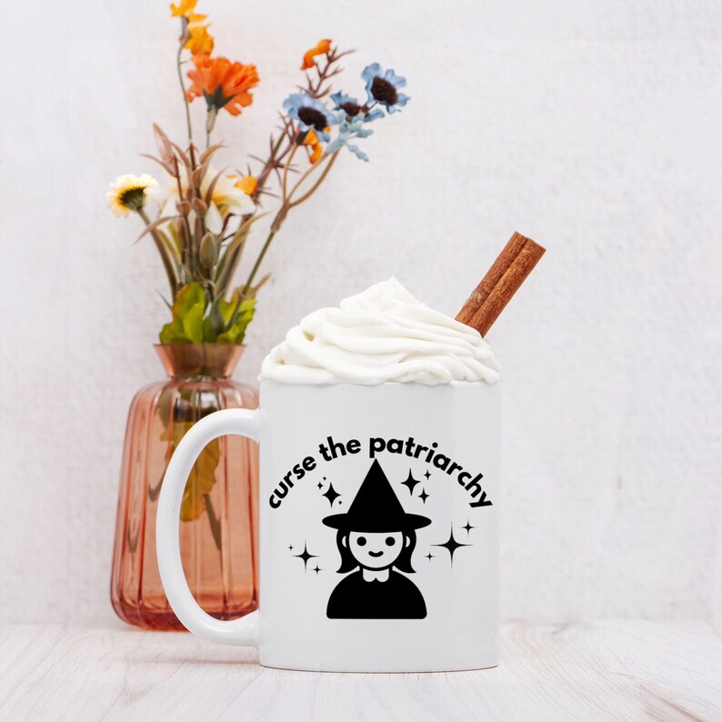 May include: White ceramic mug with a black silhouette of a witch wearing a pointed hat and the text "curse the patriarchy" with stars around the text. The mug is filled with a white beverage topped with whipped cream and a cinnamon stick.