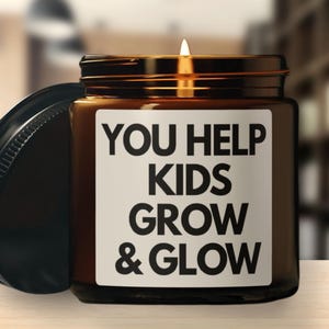 May include: A lit candle in a brown glass jar with a black lid. The jar has a white label with the text "YOU HELP KIDS GROW & GLOW" in bold black letters. The lid is off to the side.