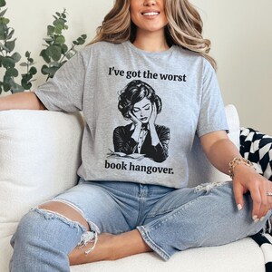 Op de afbeelding: Een grijs T-shirt met een zwart-witte illustratie van een vrouw die haar hoofd in haar handen houdt. De tekst op het shirt luidt "Ik heb de ergste kater van een boek."
