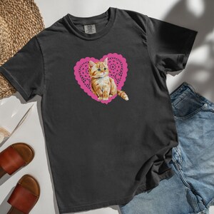 May include: A black t-shirt with a pink heart-shaped doily featuring a cute orange tabby kitten.