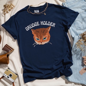 May include: A navy blue t-shirt with a cartoon image of an orange cat with a grumpy expression. The text "GRUDGE HOLDER" is printed in white above the cat.