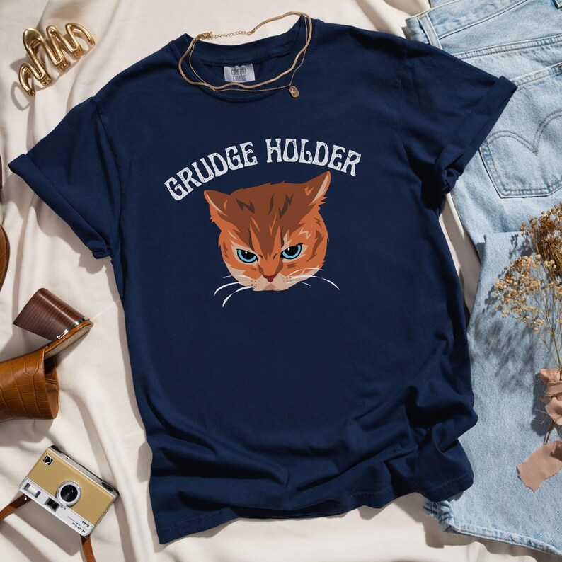 May include: A navy blue t-shirt with a cartoon image of an orange cat with a grumpy expression. The text "GRUDGE HOLDER" is printed in white above the cat.