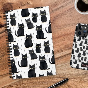 May include: A black and white spiral notebook with a black cat pattern. The notebook has a white background with black cats with yellow eyes. The notebook has a black spiral binding. The notebook is on a wooden surface. A black phone case with a black cat pattern is also on the surface. The phone case has the name "Maddie" printed on it.