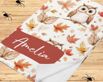 Könnte beinhalten: Eine weiße Decke mit einem herbstlichen Muster mit braunen und orangefarbenen Blättern und einer Eule, die auf einem Ast sitzt. Die Decke hat ein rotes Rechteck mit dem Namen "Amelia" in weißer Schrift. Die Decke ist gefaltet.