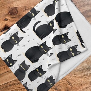 May include: White fleece blanket with a black and white repeating pattern of black cats with yellow eyes. The blanket is folded in half, showing the white fleece backing.