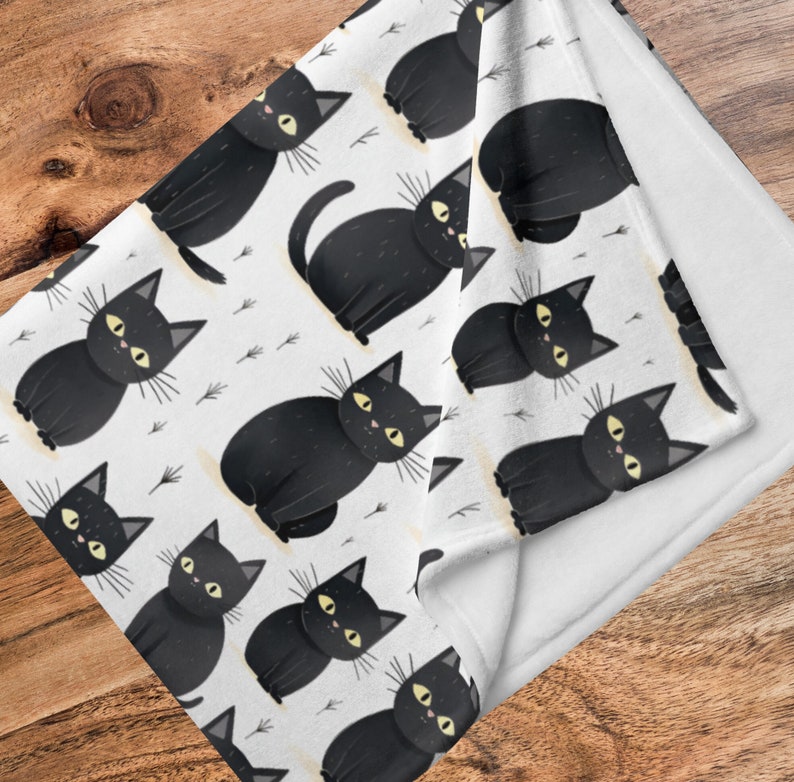 May include: White fleece blanket with a black and white repeating pattern of black cats with yellow eyes. The blanket is folded in half, showing the white fleece backing.