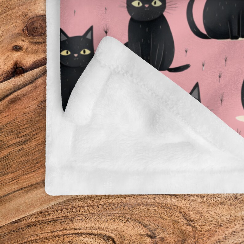 以下が含まれることがあります： ピンクと黒のフリースブランケットで、黒い猫が繰り返し描かれたパターンになっています。ブランケットは半分に折り畳まれており、柔らかく白いフリースの裏地が見えています。