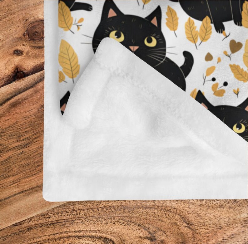 May include: White fleece blanket with a black cat and yellow leaf pattern. The blanket is folded in half and the pattern is visible on the front side.