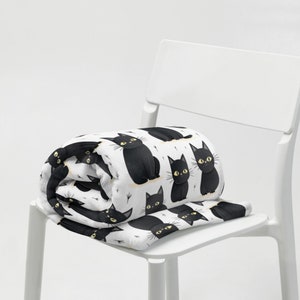 May include: A white fleece blanket with a black cat pattern. The blanket is folded and placed on a white chair.