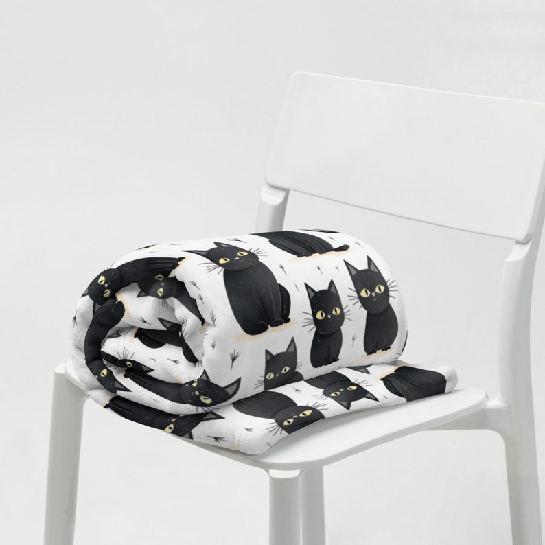 May include: A white fleece blanket with a black cat pattern. The blanket is folded and placed on a white chair.