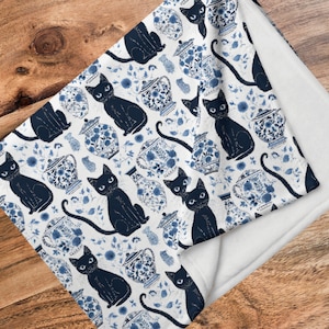 K&ouml;nnte beinhalten: Wei&szlig;e Fleece-Decke mit einem schwarzen Katzenmotiv und einem sich wiederholenden Muster aus blauen und wei&szlig;en Blumen.