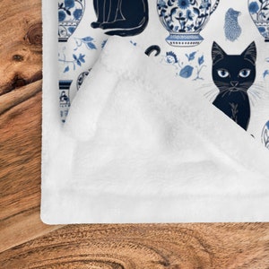 K&ouml;nnte beinhalten: Wei&szlig;e Fleece-Decke mit einem sich wiederholenden Muster aus schwarzen Katzen und blau-wei&szlig;en Porzellanvasen. Das Muster erinnert an traditionelles chinesisches Blau-Wei&szlig;-Porzellan.