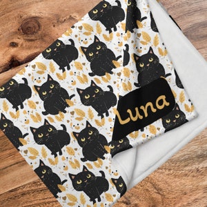 May include: A white fleece blanket with a black and gold print of cartoon cats. The blanket has a black tag with the name "Luna" written in yellow.