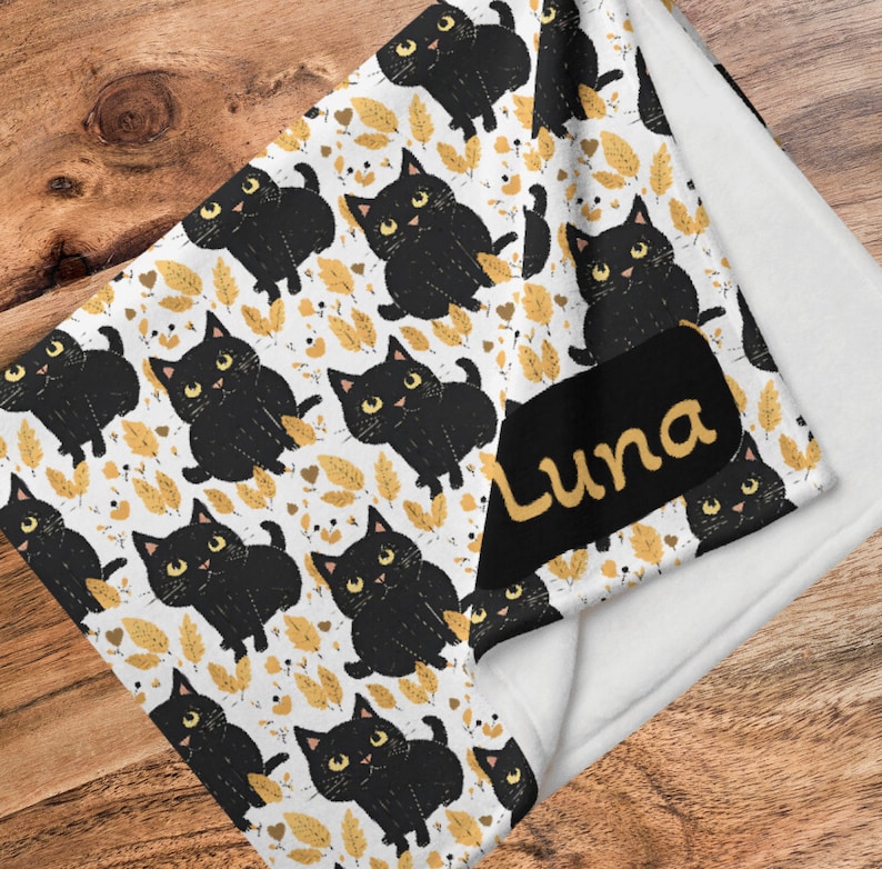 May include: A white fleece blanket with a black and gold print of cartoon cats. The blanket has a black tag with the name "Luna" written in yellow.