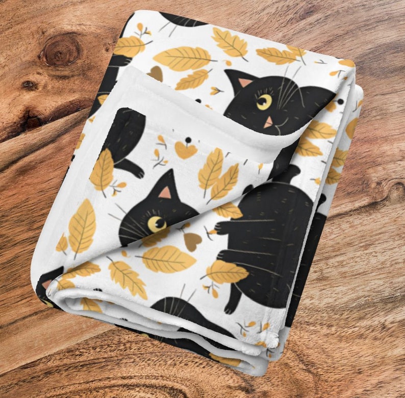 May include: A white fleece blanket with a black cat and yellow leaves pattern. The cat is depicted in various poses, including sitting, standing, and lying down. The leaves are scattered throughout the blanket, creating a whimsical autumnal design.