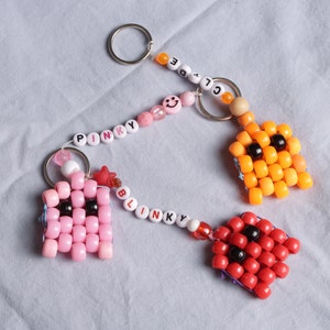 May include: Two Pac-Man character keychains made of colorful beads. The pink keychain has the word "PINKY" spelled out in white beads and the red keychain has the word "BLINKY" spelled out in white beads.